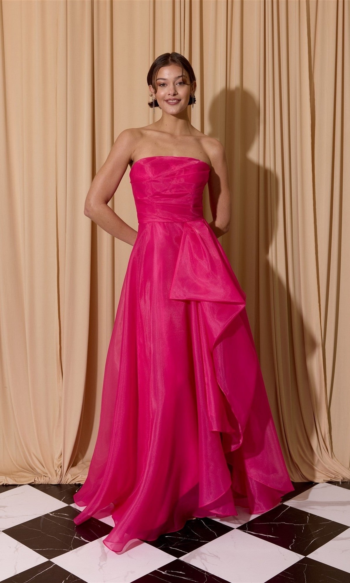 A woman stands on a black-and-white checkered floor in front of beige curtains, wearing the Eureka EK206 Long Prom Dress in fuschia.
